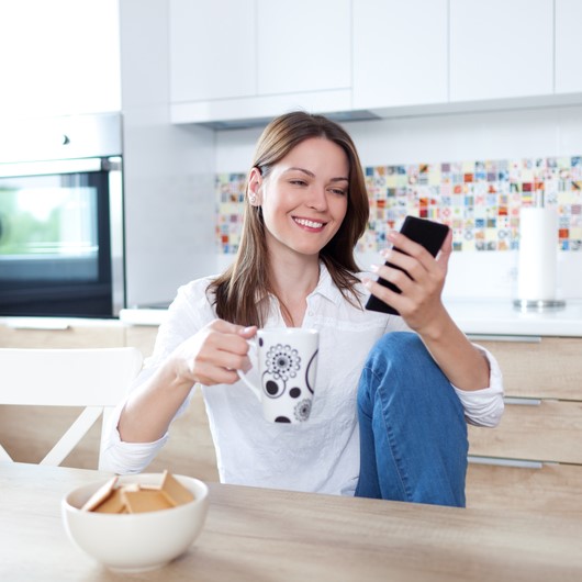 woman seated at a kitchen table holding a mug and smiling while on her phone